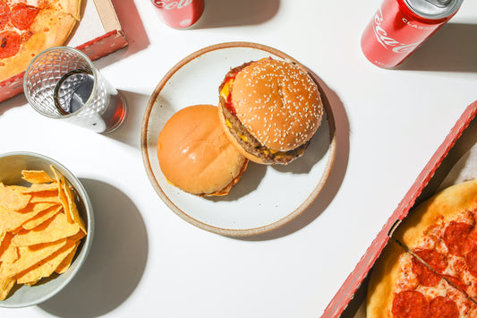 Table filled with various junk foods such as burgers, chips, soda, and pizza